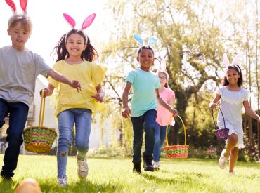 Group Of Children Wearing Bunny Ears Running To Pick Up Chocolate Egg On Easter Egg Hunt In Garden