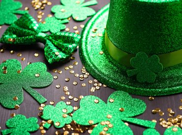 Happy Saint Patrick's Day greeting card with traditional symbols, shamrock, green attire. Green hat, bow tie, St Patricks Day shamrocks, golden confetti on wooden background, copy space, top view